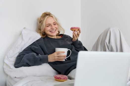 Lifestyle and people concept. Happy blond woman lying in bed with food, drinking tea and eating doughnut, resting at home, spending time alone, watching tv show.