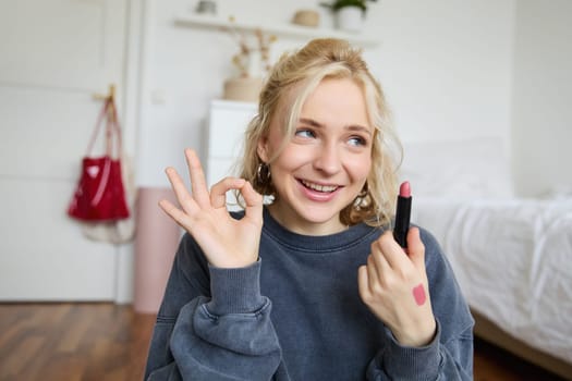 Young female blogger, content creator showing lipstick and okay hand sign, recommending beauty product for her audience on social media, recording vlog in room.