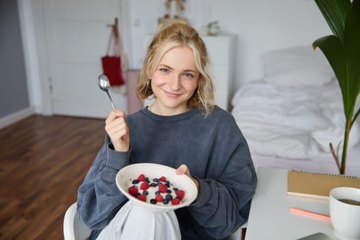 Portrait of cute smiling blond woman eats breakfast in her bedroom, looking at camera, holding bowl with dessert and spoon.