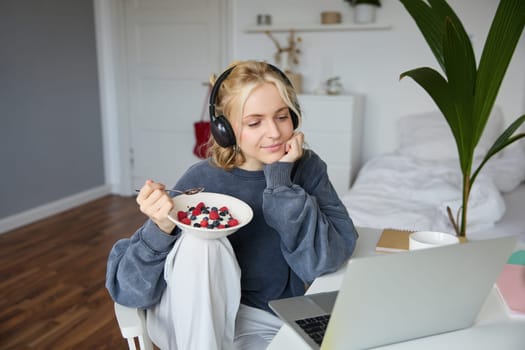 Portrait of happy young blond woman, sitting in a room, watching movie on laptop and eating healthy breakfast, drinking tea, resting on weekend.