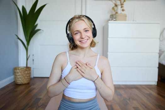 Portrait of woman feeling relaxed and in peace after meditation or yoga training at home, holding hands on chest, wearing headphones, smiling at camera.