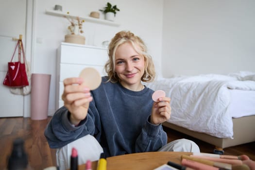 Portrait of young woman, content creator, showing beauty makeup products at camera, sitting on floor in bedroom and smiling.