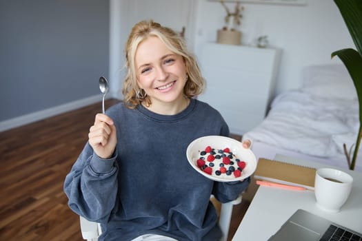 Portrait of smiling girl vlogger, blogger holding bowl with dessert, showing it on camera to audience, recording vlog in room.