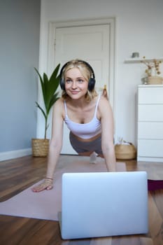 Vertical shot of young blond woman at home, wearing wireless headphones, looking at laptop, standing in plank, watching fitness tutorial online.