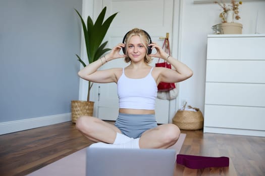 Portrait of young fitness girl, woman workout at home, listens to fitness instructor in wireless headphones, follows exercises on laptop.