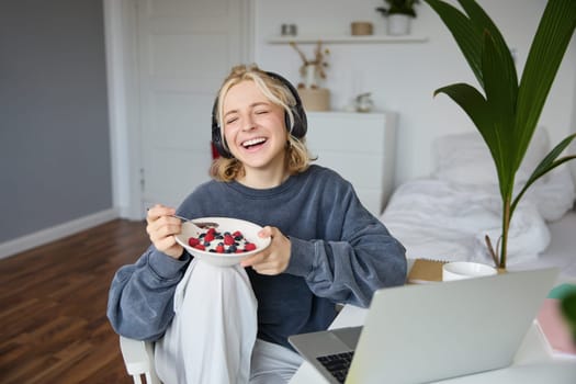 Portrait of happy young blond woman, sitting in a room, watching movie on laptop and eating healthy breakfast, drinking tea, resting on weekend.