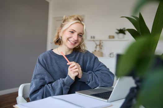 Portrait of young smiling woman, student talking to teacher online via video chat, doing course, attends remote course in internet, making notes.