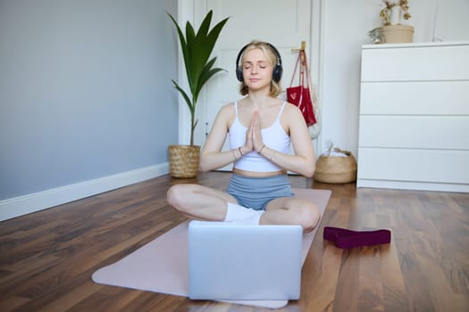 Portrait of young, relaxed woman in headphones, listens to meditation music on laptop, practice yoga on rubber mat, holding hands clasped together in namaste sign.