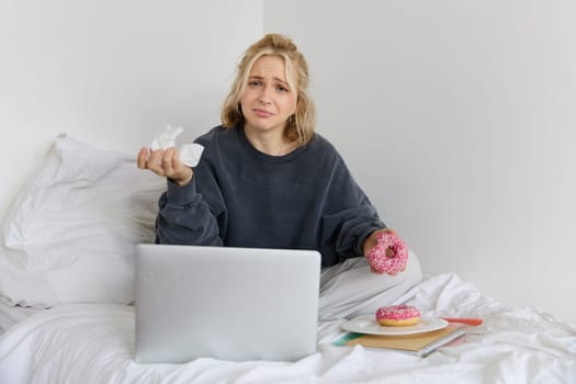 Portrait of disappointed, depressed young woman, crying, sitting on bed with laptop, eating comfort food, holding doughnut and wiping tears off.