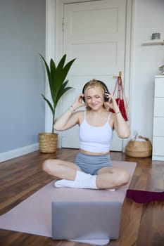 Portrait of young woman at home, connects to online gym training session, following fitness instructions on laptop, wearing wireless headphones during workout.