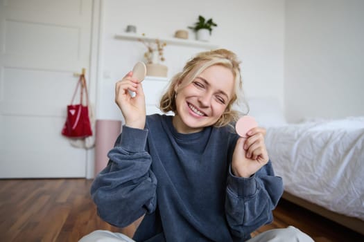 Portrait of young woman, content creator, showing beauty makeup products at camera, sitting on floor in bedroom and smiling.