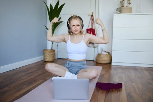 Portrait of young athletic woman doing workout at home, shows her muscles, strong biceps, sits on yoga mat and wears wireless headphones.