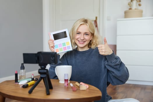 Portrait of young smiling woman, girl reviewing palette eyeshadow, shows thumbs up, recording video blog, vlogging in her room, recommending makeup product.