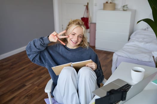 Portrait of smiling cute woman, lifestyle blogger, sits in her room with daily journal or planner, records video on digital camera, creates content for social media about daily routine.