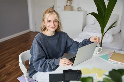 Portrait of woman sitting at desk with laptop, recording video of herself on laptop, making lifestyle video for social media account.
