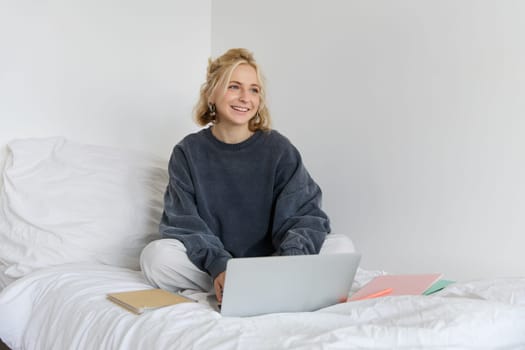 Image of happy young woman, student e-learning from home, connect to online course on her laptop, sits on bed with notebooks, smiling and looking happy.