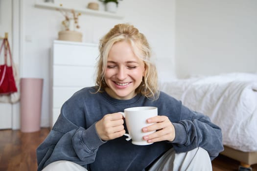 Portrait of young blond woman in casual clothes, sits on bedroom floor with cup of tea, drinking and looking at camera.