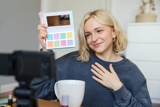 Lifestyle blogger, records video in her room, has a camera on coffee table, shows eyeshadow palette to her followers, does makeup tutorial, vlogger working indoors, creates content for social media.