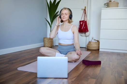 Portrait of fit and healthy woman at home, practice yoga, sitting on rubber mat, listening to instructions online, using meditation music to relax, following guidance on laptop.