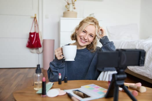 Portrait of blond young lifestyle blogger, woman records video of her talking about life and beauty, sits in front of camera, holds cup, drinks tea, does blog content for social media account.