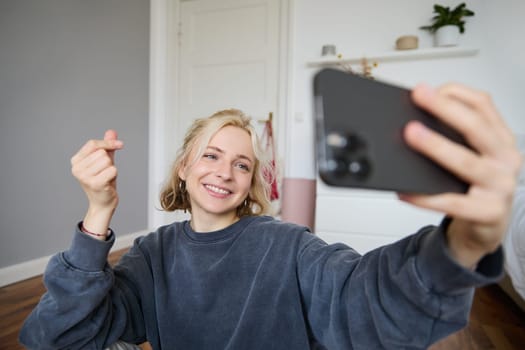 Portrait of cute smiling young woman with smartphone, taking selfies in her room, showing heart sign, taking pictures or video chatting.