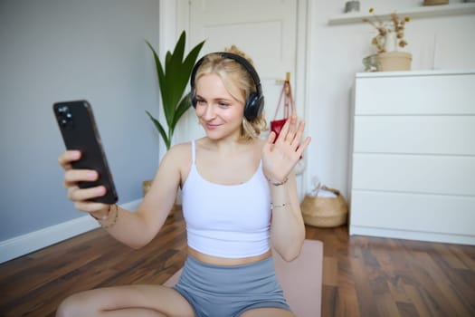 Portrait of young sporty woman in headphones, records video on her smartphone, live streaming and saying hello to followers while doing workout training at home.