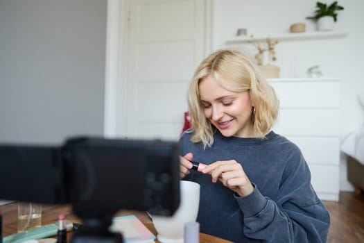 Close up portrait of smiling young woman with video camera in her room, showing mascara, reviewing beauty products for social media account. Beauty blogger creates vlog about makeup.