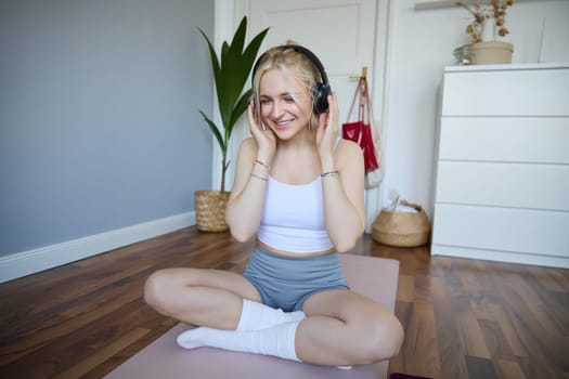 Sport and wellbeing concept. Smiling woman in wireless headphones, sitting on yoga mat, listens to music and workout at home, enjoys fitness training session.