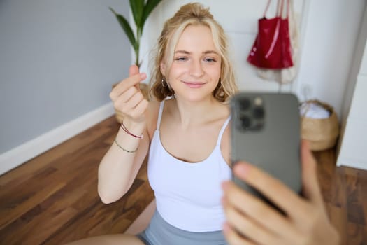 Portrait of young beautiful blond woman, fitness instructor doing exercises at home on yoga mat, taking selfies on smartphone, recording video of herself for social media account about workout.