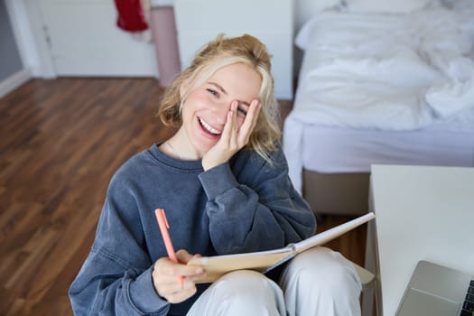 Portrait of happy, young blond woman, sitting in bedroom with notebook and pen, laughing and smiling, writing in journal, doing homework and smiling.