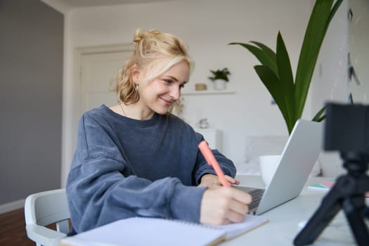 Portrait of smiling blond woman writing in notebook, making notes, recording content for social medial on digital camera, looking at laptop, working or studying.