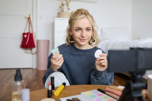 Portrait of young beauty blogger, woman gives advices to audience, how to apply makeup, sits in front of digital camera, holds foundation cream and cotton pad.