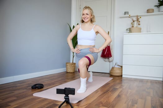 Portrait of young athletic woman, vlogger making a video about home workout, fitness instructor shooting how to do leg exercises, using elastic resistance band, using yoga mat.