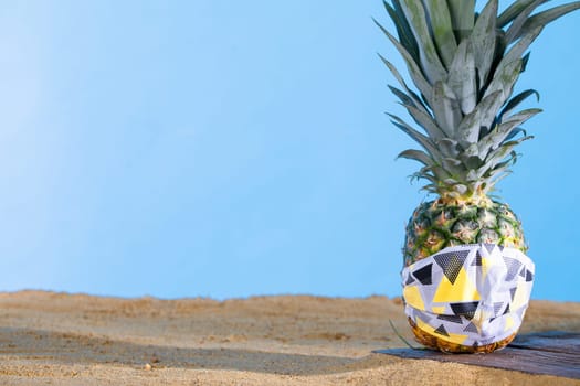 Pineapple in a protective mask stands on the sandy sea beach.