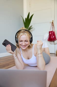 Vertical shot of young fitness woman in headphones, lying on rubber mat, relaxing after workout training session, holding smartphone, smiling at camera. Lifestyle and wellbeing concept