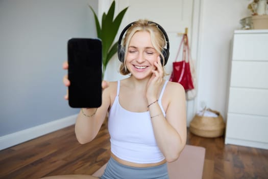 Portrait of smiling, beautiful young woman, showing her smartphone screen, wearing headphones, looking satisfied with sound quality.