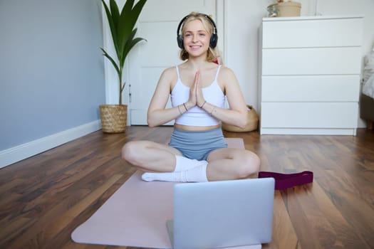 Portrait of young, relaxed woman in headphones, listens to meditation music on laptop, practice yoga on rubber mat, holding hands clasped together in namaste sign.