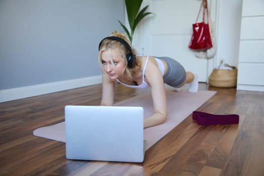 Portrait of young sporty woman doing workout, looking at fitness video on laptop in wireless headphones, standing in plank on yoga mat, following exercise tutorial.