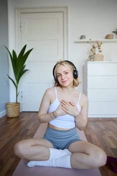 Portrait of young blond woman in headphones, feeling calm and relaxed after meditation, yoga training, sitting on yoga mat in her room. Wellbeing, sport and people concept