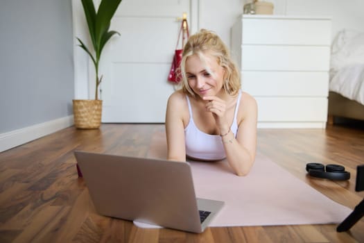 Portrait of beautiful blond woman looking at fitness video tutorials on laptop, lying on rubber yoga mat, following workout instructions online.