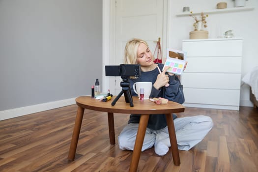 Portrait of beautiful lifestyle blogger, girl records a video on her camera for social media, shows palette of eyeshadows, does a makeup tutorial for her followers, sits in her room.