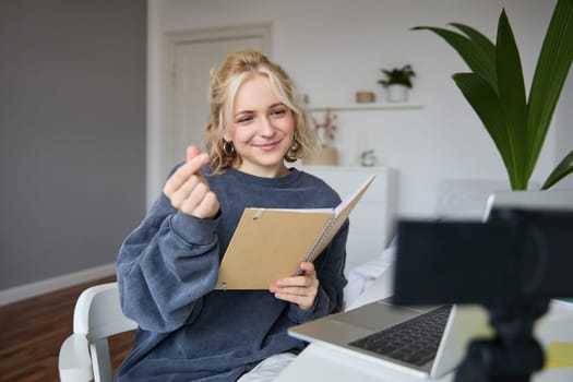 Beautiful young woman in her room, sits indoors, records video on digital camera, holds notebook and her notes, shows heart finger sign.