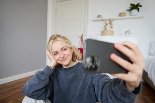 Portrait of young stylish girl sits on bedroom floor, takes selfies on her smartphone, posing for photo on social media app, smiling and looking happy at camera.