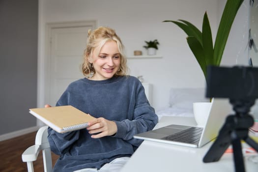Young smiling blond woman, sits near laptop, uses digital camera to record video blog, creates lifestyle content for social media, records tutorial, shows notebook, reads her notes.