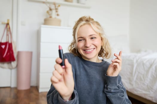 Portrait of young social media influencer, woman recording a video with beauty products, showing her makeup on camera, holding lipstick and smiling, sitting on floor.