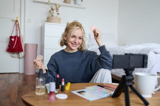 Portrait of young woman, beauty content creator, sitting in a room in front of digital camera, recording makeup tutorial vlog, showing cosmetic facial products.