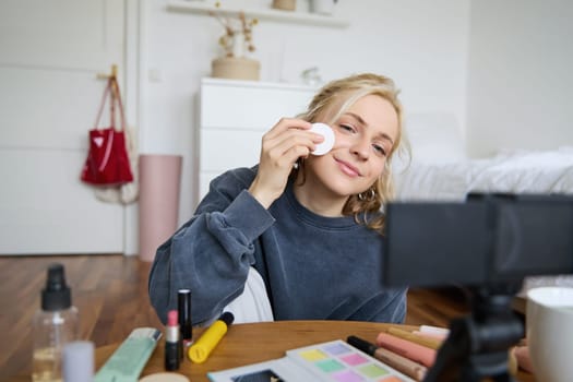 Portrait of smiling young woman applies makeup, shows beauty tutorial on video, records vlog on digital camera in her room, recommends cosmetic products.