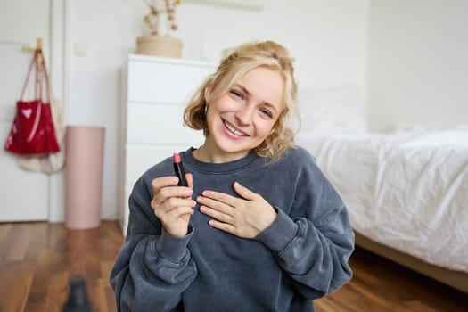 Portrait of cute, charismatic beauty blogger, woman sits in a room with lipstick in hand, talking about makeup, chatting with followers, recording online stream on social media app.