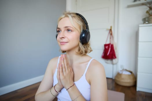 Close up portrait of young relaxed woman in headphones, holding hands together in namaste gesture, listening to meditation podcast, practice yoga.