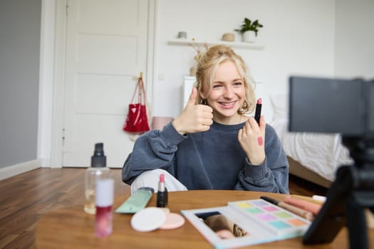 Happy young content creator, woman showing lipstick, recording lifestyle, beauty vlog on digital camera, makes thumbs up hand gesture, recommending makeup product.